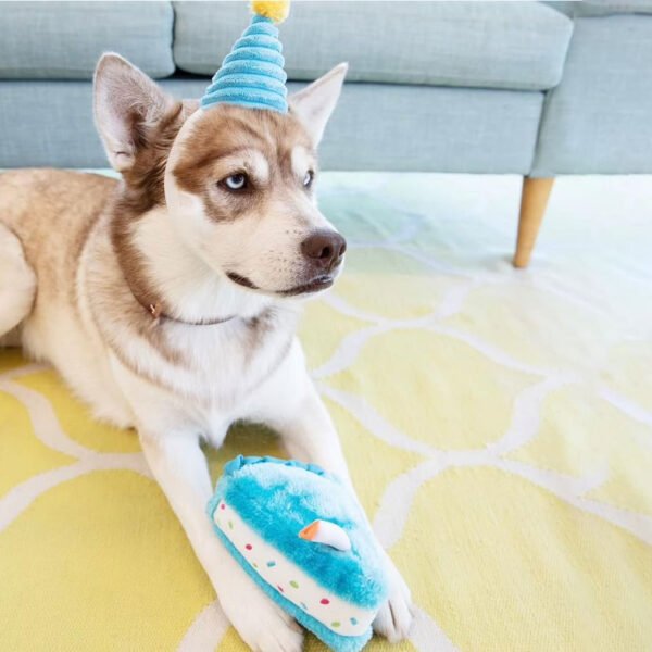 Dog lying on a yellow and white patterned rug with its head resting on a blue plush toy, with a grey sofa in the background.