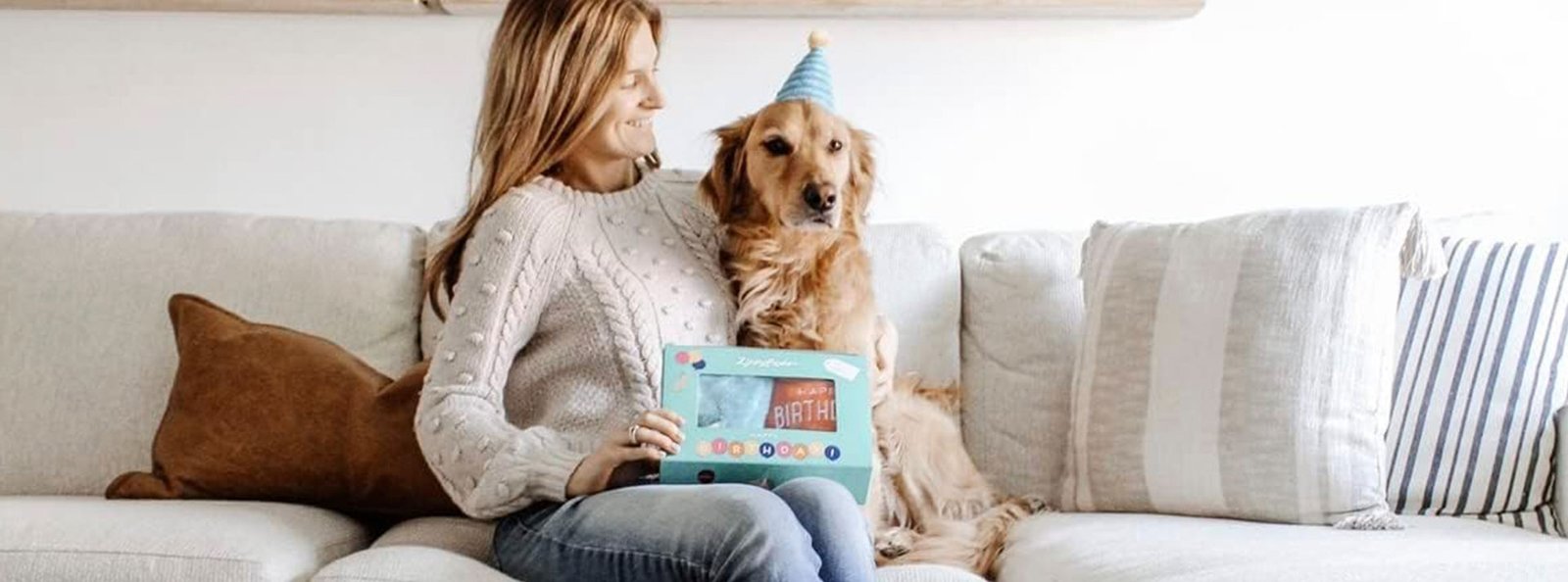 Golden retriever wearing a blue birthday hat, sitting beside a person on a couch, both looking at a birthday card.