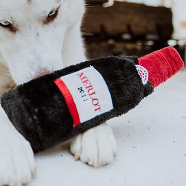 White dog holding a plush squeaky toy resembling a Merlot wine bottle, labeled “MERLOT” with ZippyPaws branding, designed for pet playtime.