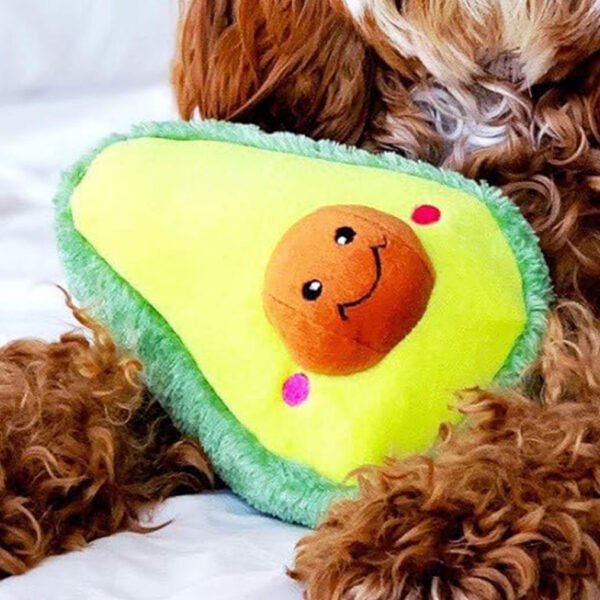 Plush avocado-shaped dog toy with a happy face, nestled between the paws of a fluffy brown dog on a white background