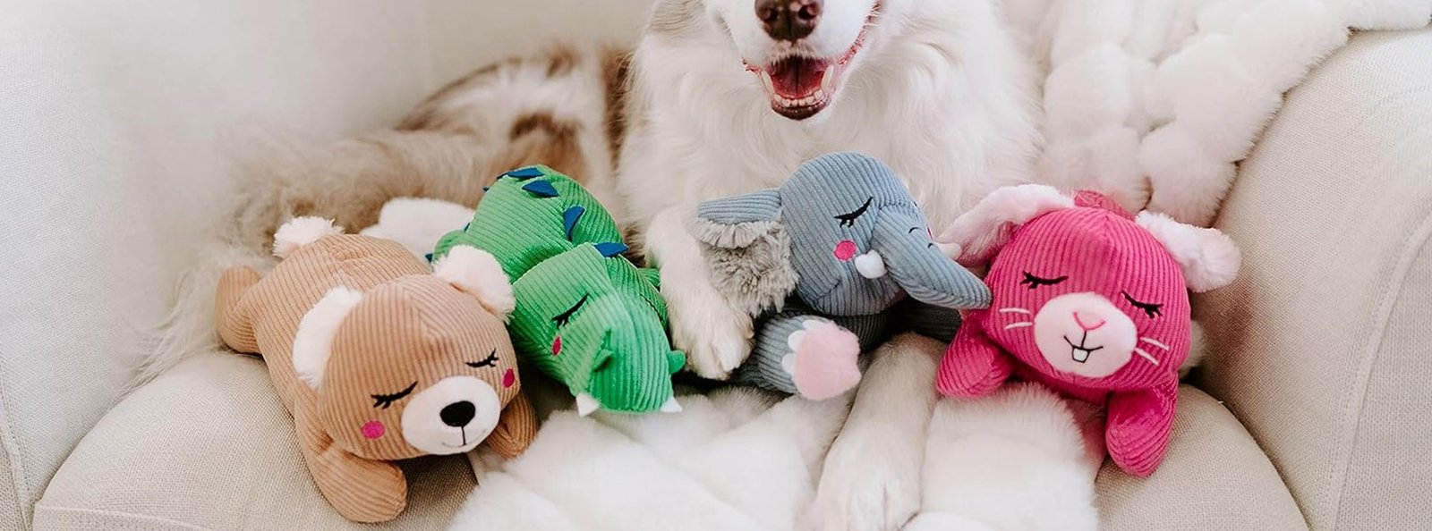 Joyful dog surrounded by colorful Snoozies plush toys on a soft white blanket