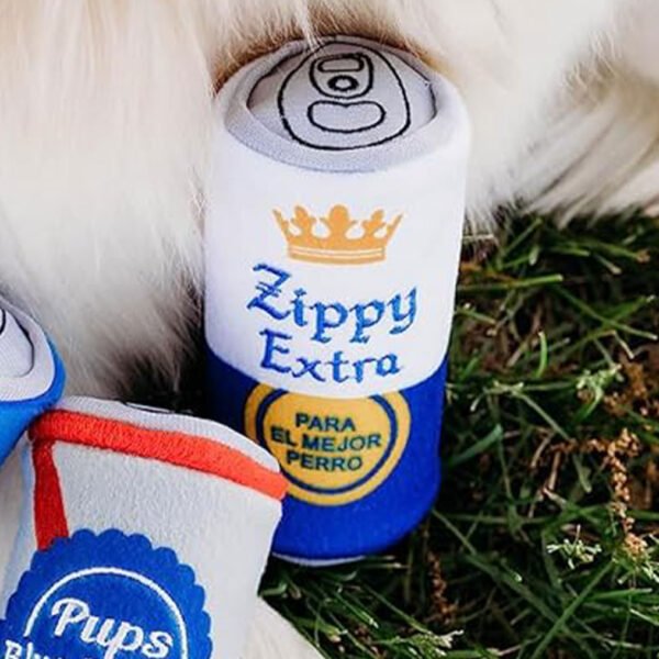 Close-up of a plush dog toy resembling a can, with “Zippy Extra” in bold letters, nestled in the grass beside another toy