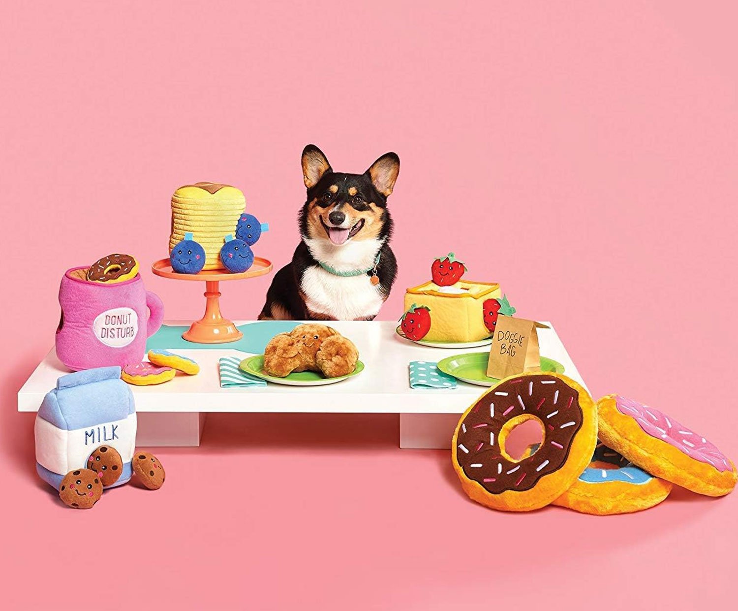 A variety of colorful plush toys, including food-shaped items and a blue toy bone, arranged on a white table with a black and tan dog sitting in the center, against a pink background