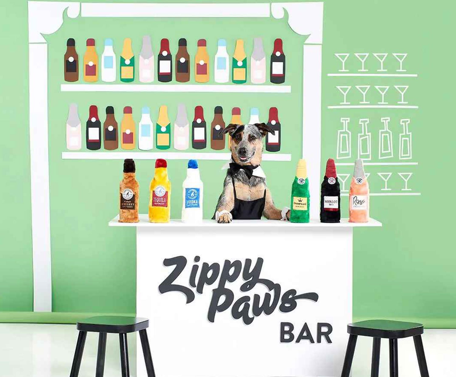 Dog standing behind a bar counter labeled “Zippy Paws Bar,” wearing a black bow tie and surrounded by plush toy bottles, symbolizing the joy and comfort of pet toys.