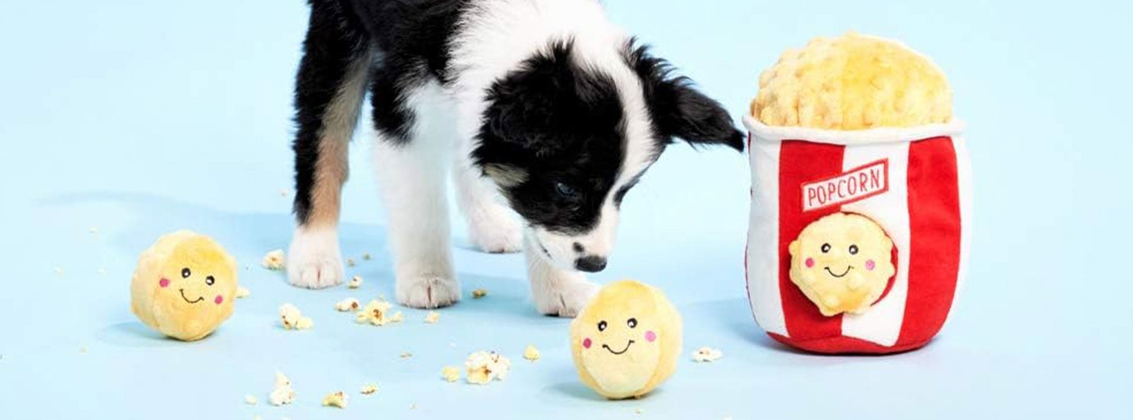 A black and white puppy sniffing a full popcorn bag with a smiley face, surrounded by scattered popcorn and two smiley-faced balls on a light blue background