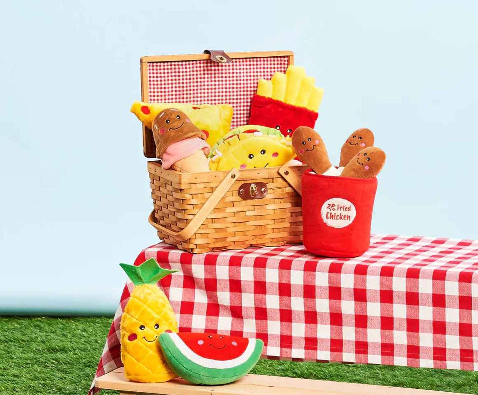 An assortment of the squeaky plush zippypaws dog toys nomnomz collection, including the watermelon, pineapple, pizza slice, ice cream, fries and a red ‘Fried Chicken’ bucket toy, resting on a classic red and white checkered picnic blanket over fresh green grass.