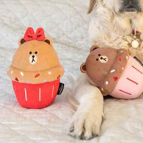 “A plush squeaker dog toy designed to resemble a teddy bear cupcake, with a tan bear top featuring a cute face and red bow, sitting in a red cupcake wrapper, next to another similar toy on a quilted background.