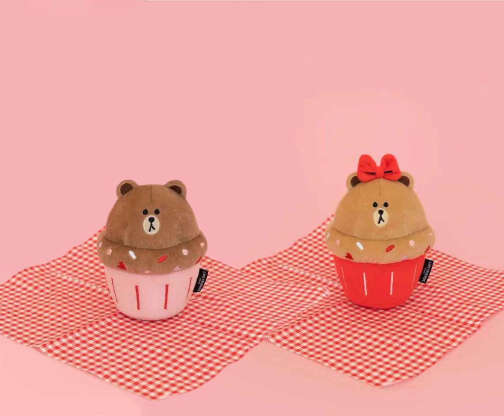 Two NomNomz® Cupcake plush squeaker dog toys, one with a bear face on a pink background and the other with a bow on a red background, both resting on a checkered cloth.