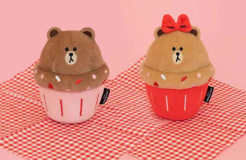 Two NomNomz® Cupcake plush squeaker dog toys, one with a bear face on a pink background and the other with a bow on a red background, both resting on a checkered cloth.