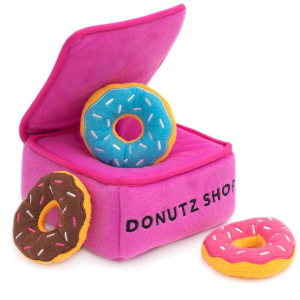 A vibrant pink plush ‘Donutz Shop’ box with a lid open to reveal one blue frosted donut toy inside, and two additional donut toys with pink and chocolate frosting beside the box.