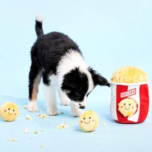 A black and white puppy interacts with a ZippyPaws Popcorn Bucket dog toy, accompanied by three plush popcorn-shaped squeaky toys scattered around.