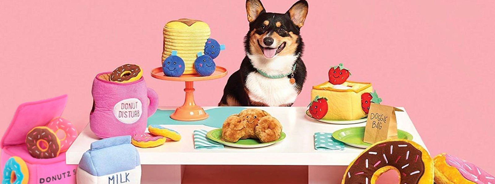A cheerful Corgi sits behind a colorful array of dog toys resembling human treats on a pink background, promoting fun and engagement for pets.