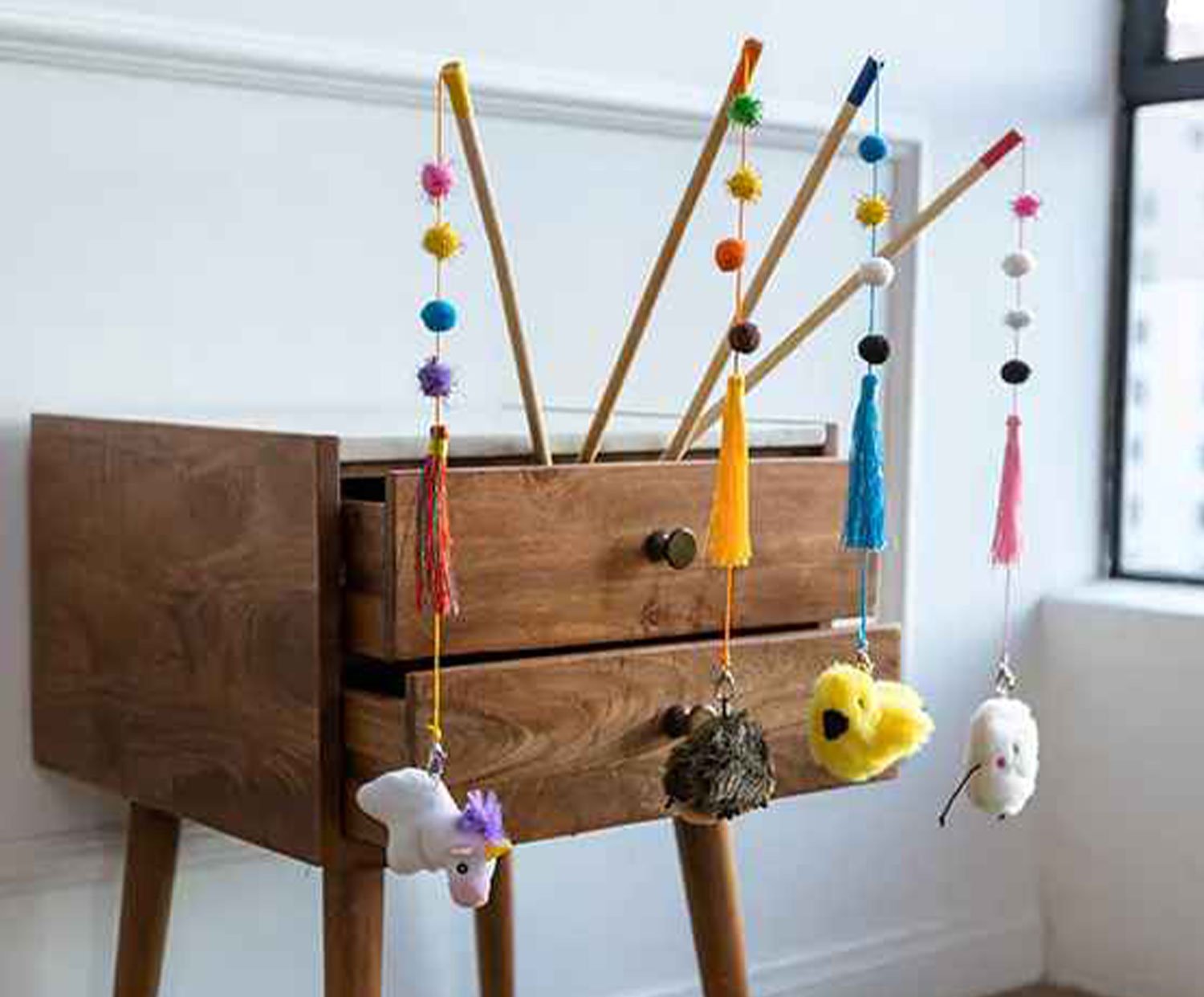 Various cat wand toys with colorful beads, ribbons, and plush toys like a unicorn, bird, and mouse displayed on a wooden side table.