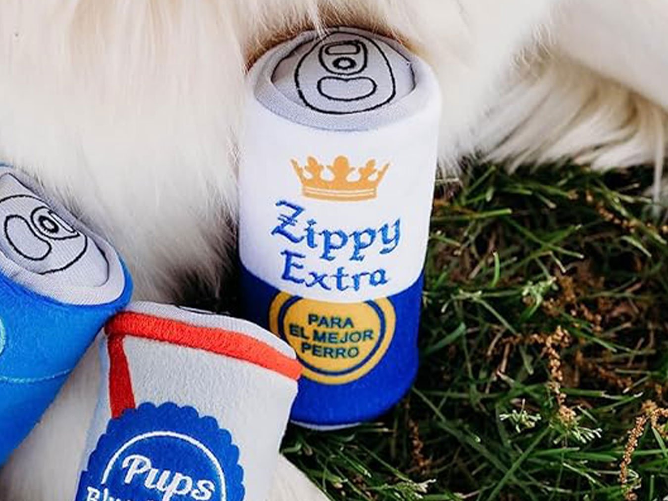 Close-up of a plush dog toy resembling a can, with “Zippy Extra” in bold letters, nestled in the grass beside another toy