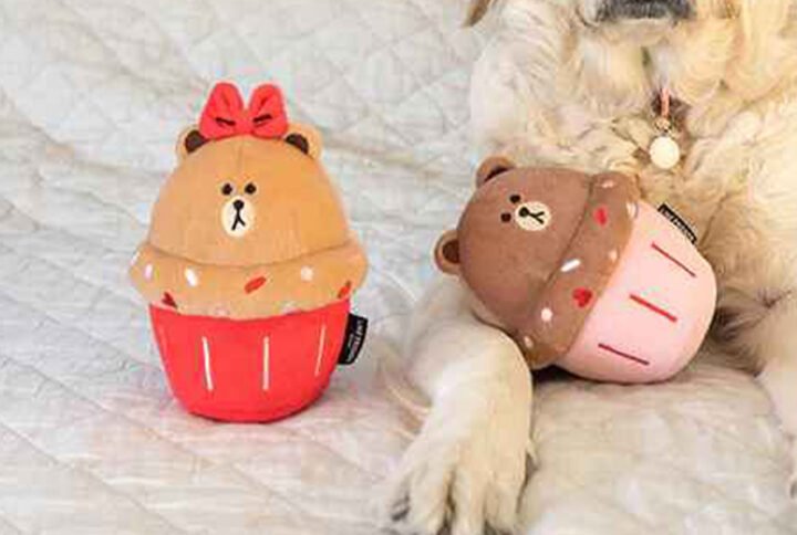 “A plush squeaker dog toy designed to resemble a teddy bear cupcake, with a tan bear top featuring a cute face and red bow, sitting in a red cupcake wrapper, next to another similar toy on a quilted background.