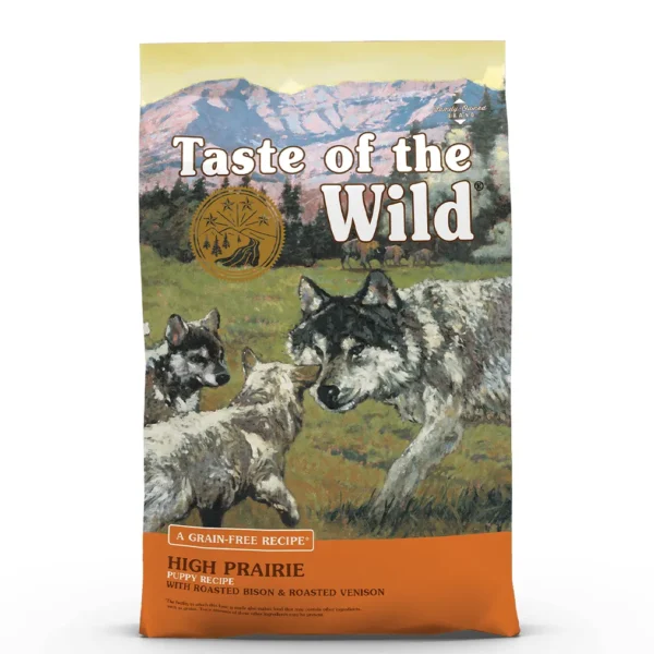Studio shot of the front of the Taste of the Wild High Prairie Puppy Recipe bag. The predominantly warm, earthy brown/orange bag features a wilderness illustration with a mountain range and pine trees in the background. In the mid-ground, a herd of bison is visible on a prairie with an alert adult wolf accompanied by two wolf pups in the foreground. Key texts include "HIGH PRAIRIE PUPPY RECIPE", "WITH ROASTED BISON & ROASTED VENISON", and "A GRAIN-FREE RECIPE", with a gold circular emblem above the illustration and small print indicating "Family‑Owned BRAND" plus a disclaimer.