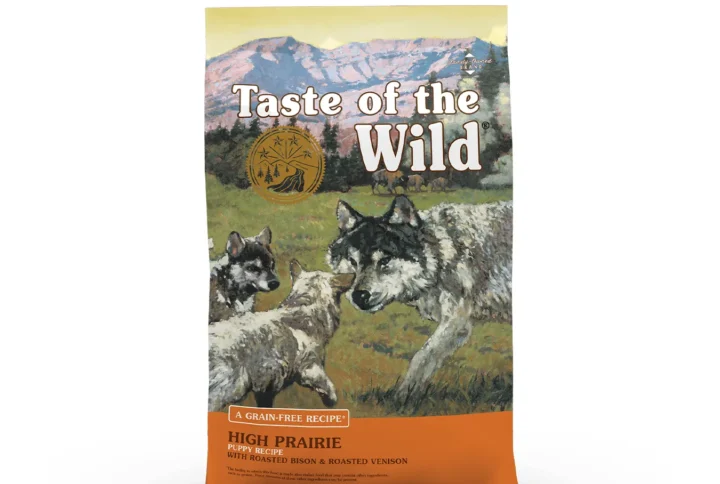 Studio shot of the front of the Taste of the Wild High Prairie Puppy Recipe bag. The predominantly warm, earthy brown/orange bag features a wilderness illustration with a mountain range and pine trees in the background. In the mid-ground, a herd of bison is visible on a prairie with an alert adult wolf accompanied by two wolf pups in the foreground. Key texts include "HIGH PRAIRIE PUPPY RECIPE", "WITH ROASTED BISON & ROASTED VENISON", and "A GRAIN-FREE RECIPE", with a gold circular emblem above the illustration and small print indicating "Family‑Owned BRAND" plus a disclaimer.