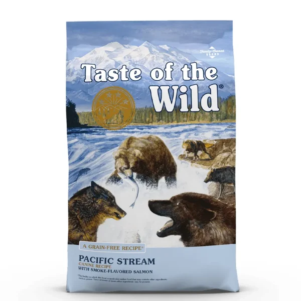 Studio shot of the front of the Taste of the Wild Pacific Stream Canine Recipe bag. The predominantly blue bag features a dramatic wilderness scene with snow-capped mountains, a forest, and a rushing river. In the foreground, a large grizzly bear catches a salmon while a smaller bear and a wolf watch from the riverbanks. Key text includes "PACIFIC STREAM CANINE RECIPE," "WITH SMOKE-FLAVORED SALMON," and "A GRAIN-FREE RECIPE," with a gold circular emblem and small print details like "DIAMOND PET FOODS" and production disclaimers.