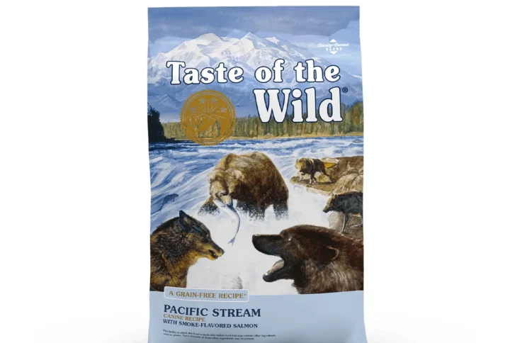 Studio shot of the front of the Taste of the Wild Pacific Stream Canine Recipe bag. The predominantly blue bag features a dramatic wilderness scene with snow-capped mountains, a forest, and a rushing river. In the foreground, a large grizzly bear catches a salmon while a smaller bear and a wolf watch from the riverbanks. Key text includes "PACIFIC STREAM CANINE RECIPE," "WITH SMOKE-FLAVORED SALMON," and "A GRAIN-FREE RECIPE," with a gold circular emblem and small print details like "DIAMOND PET FOODS" and production disclaimers.