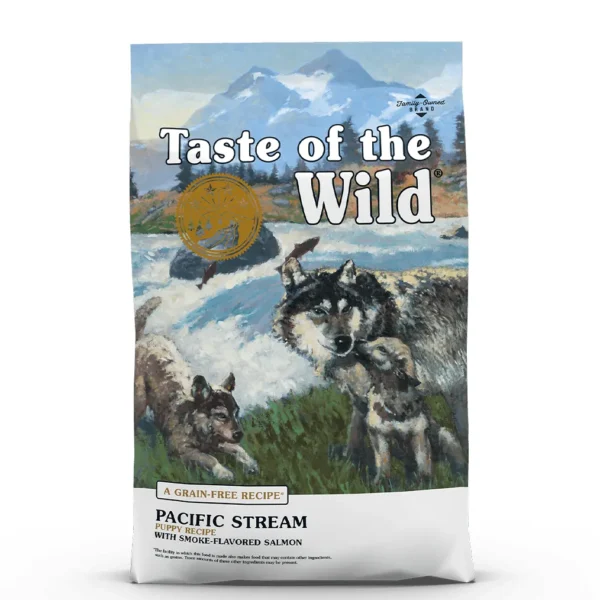 Studio shot of the Taste of the Wild Pacific Stream Puppy Recipe bag. The predominantly blue bag features a dramatic wilderness scene with snow-capped mountains, a rushing river, and an illustration of an adult wolf with two playful wolf pups. Key texts include "PACIFIC STREAM PUPPY RECIPE," "WITH SMOKE-FLAVORED SALMON," and "A GRAIN-FREE RECIPE" along with a gold circular emblem. Small print indicates "Family-Owned BRAND" and a production disclaimer.